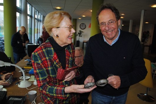 Een bezoeker van het Repair Café Schiedam is tevreden met het nieuwe onderdeel, waardoor ze haar Senseo-koffiezetapparaat weer kan gebruiken, rechts coördinator Jan Bostelaar